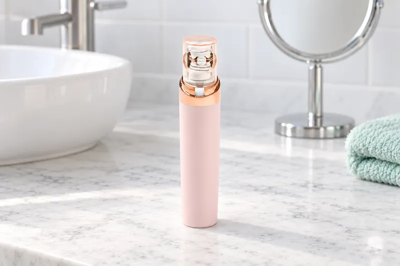 A pale pink pen-style facial epilator with a rose-gold top standing beside its clear cap, a round mirror behind
